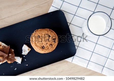 top view of a black plate on which lies a biscuit and black with white chocolate with nuts near which stands a glass of milk on a wooden table with a white napkin
