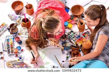 Authentic artist children girl paints with set palette watercolor paints palette and brush in morning sunlight. Painting in studio on floor. Older sister teaches younger to draw still lifes.