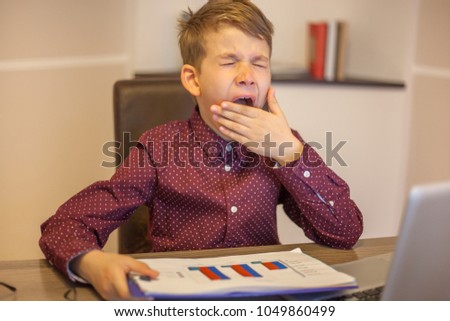 Adorable kid boss sitting at desk holding graphics and yawning