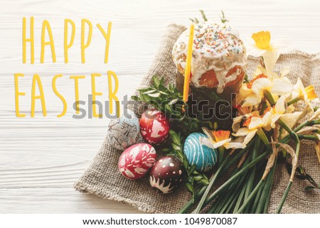 happy easter text. season's greetings card. stylish painted eggs and easter cake on white rustic wooden background with spring flowers and candle.  modern easter image