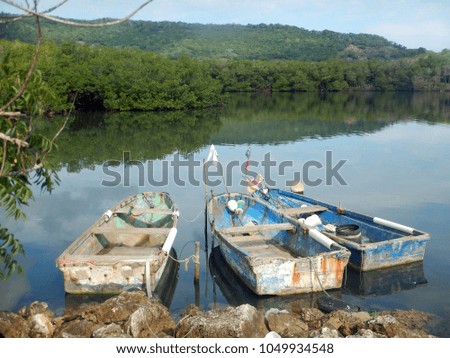 Group of three old wooden Dominican Republic fishing boats in sun bleached Caribbean colors with floats and flags staked to a rocky coast of deep blue water with lush green hills in the background.