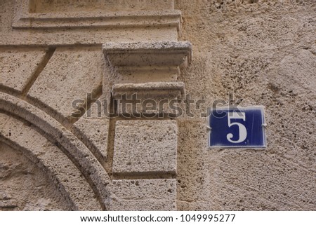 Close up outdoor front view of the number five written in white on a blue rectangular metallic plate. Symbol indicating the address in a french street. Old beige wall made of decorative stone blocks.