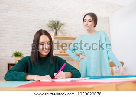 Attractive young architect wearing knitted sweater sitting at wooden desk and working on promising project while her superior keeping eye on her, interior of modern studio on background