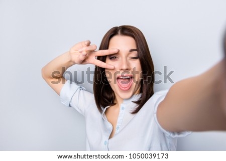 Self portrait of charming, pretty, crazy, childish, modern, stylish girl with short hair, in shirt, shooting selfie, gesture v-sign near eye, blinking with open mouth, isolated on grey background