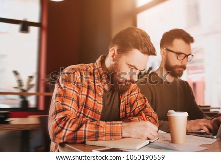 A picture of bearded guy writing down notes in his notebook while his friend is reading an article on the phone. They are very busy and concentrated.