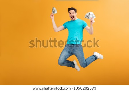 Portrait of a joyful young man in t-shirt holding bunch of money banknotes and celebrating while jumping isolated over yellow background Royalty-Free Stock Photo #1050282593