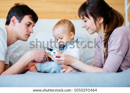 Happy family portrait. Mother, father, their son playing on bed at cozy home. Beautiful caucasian baby one years old playing with toys. Real life photo