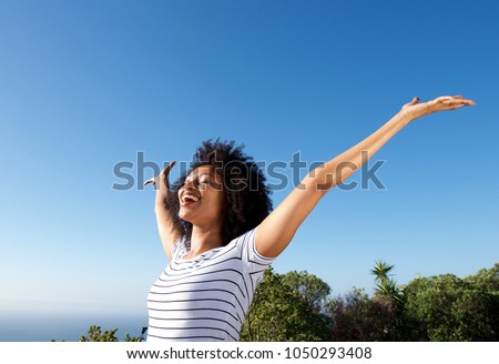 Portrait of young african woman standing outdoors with arms raised and laughing Royalty-Free Stock Photo #1050293408