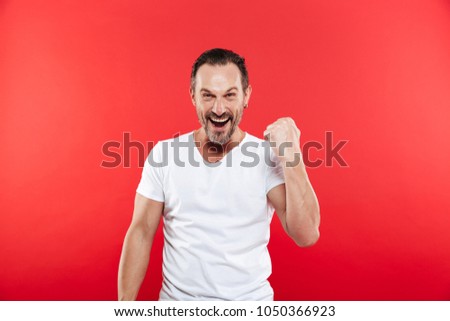 Picture of excited happy adult man standing isolated over red background looking camera make winner gesture.