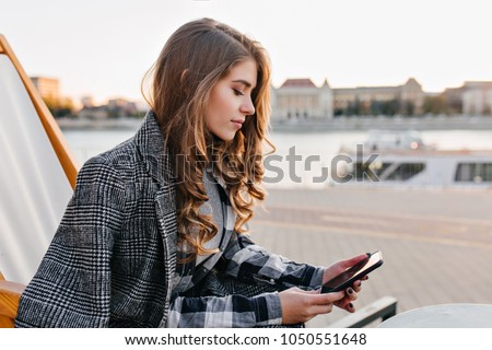 Pensive girl with curly hairstyle texting message, sitting on recliner in cold morning. Outdoor photo of thoughtfully young female model in coat chilling in chaise-longue.