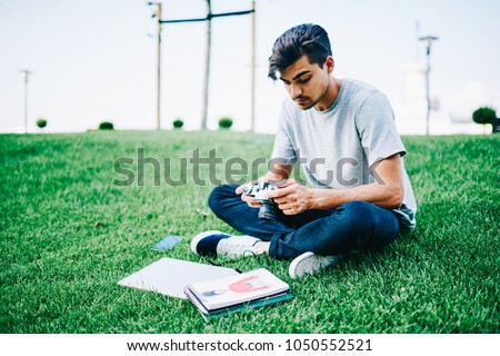Skilled student making settings on vintage camera to making photos of design sketches for studying project sitting on green grass in park.Male blogger choosing pictures for publication