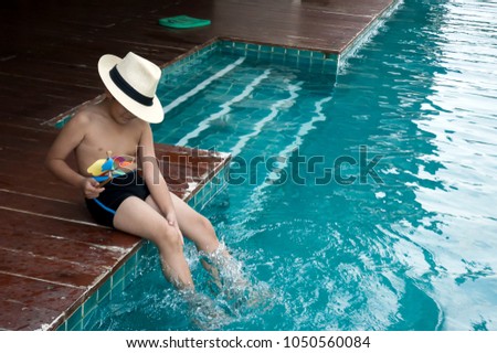 Asian handsome boy sitting at swimming pool, learning to swim, want to be swimmer, Happy learning in summer. Relax time to play colorful windmill, sitting and raise two legs down the water.