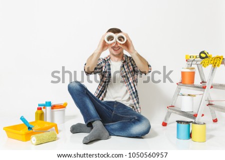 Fun male look through rolls of wallpaper as in binoculars, sit on floor, instruments for renovation apartment isolated on white background. Gluing accessories, painting tools. Repair home concept