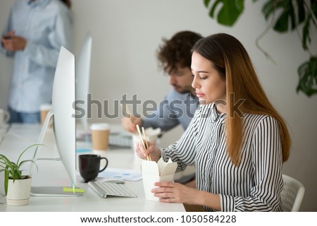 Attractive young woman enjoying chinese asian food during office lunch break, female employee holding chopsticks eating takeaway noodle box meal at workplace with colleagues in office