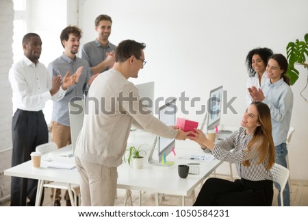 Friendly diverse corporate team congratulating happy female colleague with birthday and applauding, male employee presenting gift box greeting woman making pleasant surprise to coworker in office Royalty-Free Stock Photo #1050584231