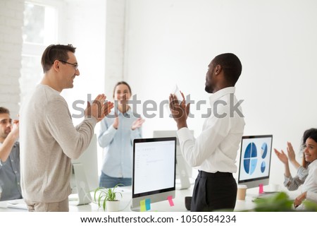 Happy motivated african american employee receiving envelope with reward or bonus from caucasian boss appreciating good work results while business team applauding supporting colleague in office Royalty-Free Stock Photo #1050584234