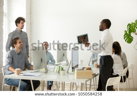 New african employee unpacking boxes and talking to friendly colleagues on first working day in office, smiling happy black worker having pleasant conversation with coworkers showing photo in frame