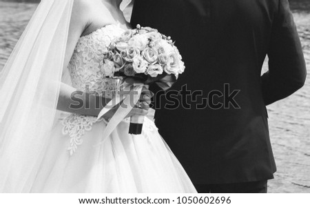 Beautiful bride in wedding satin lace dress without shoulders is hugging with her groom. Colorful bouquet with ribbon in hands. Warm embrace and love story of wedding couple. Black and white.