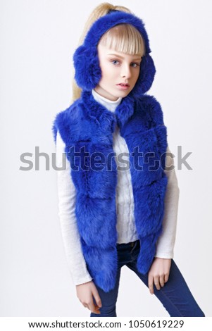 beautiful blonde teen girl dressed in blue fur vest and earmuffs posing in studio over white background