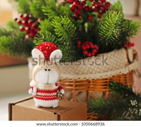 Handmade knitted toy. Christmas Bunny in a red sweater on a background of fir branches and red berries