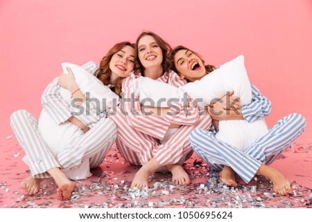 Attractive women 20s wearing leisure clothings sitting barefoot on floor with legs crossed and hugging pillows during girlish sleepover isolated over pink background Royalty-Free Stock Photo #1050695624