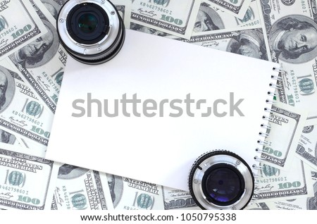 Two photographic lenses and white notebook lie on the background of a lot of dollar bills. Space for text