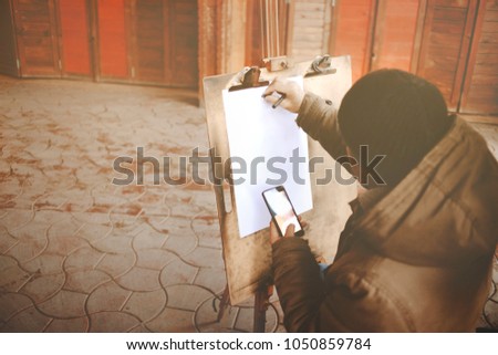 Street City artist , freelancer , working, draws a portrait of a girl , painting customers and clients with reffering to the image from phone . Copy space white blank phone and paper sheet . 