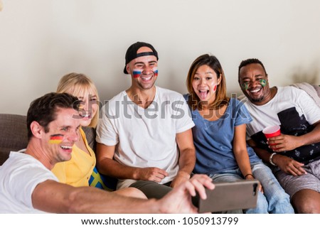 Multinational sports fans with facepainted flags laughing and taking selfie on couch