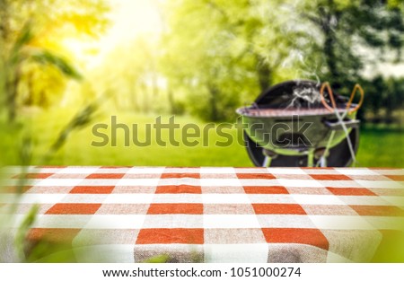 Red and white tableclothe in garden of free space for your decoration. Grill with smoke on grass.  Royalty-Free Stock Photo #1051000274