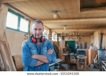 Portrait of a man who owns a small carpentry business, standing in his workshop with with arms folded and showing strong forearms, smiling confidently at the camera. 