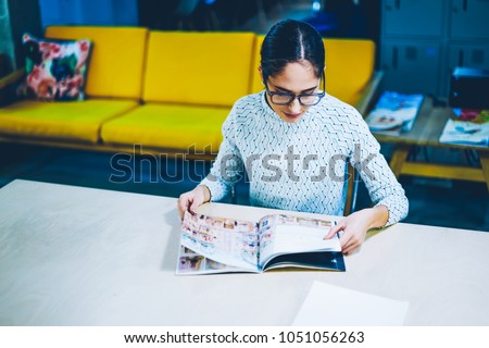 Pensive female editor of magazine reading issue for checking articles and publication while sitting at table ,concentrated woman choosing items in paper catalogue for buying furniture in office Royalty-Free Stock Photo #1051056263