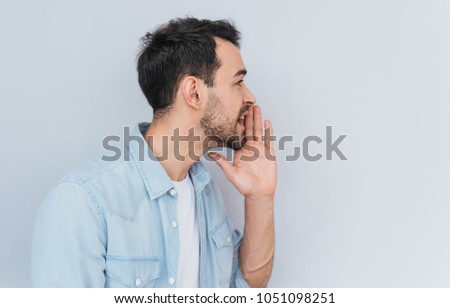 Side view portrait of attractive young Caucasian man sharing secret or whispering gossips, isolated over light grey background. Handsome male telling secret. Copy space for your advertising text. Royalty-Free Stock Photo #1051098251