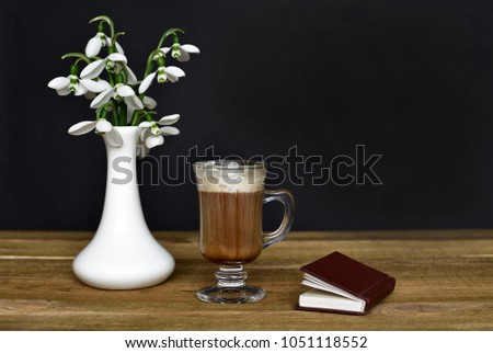 Good spring morning. Fresh snowdrops flowers bouquet in a small white vase and a coffee with leaks of creamy foam in a transparent glass and little book of poems on wooden table and black background.