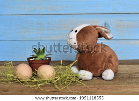 A couple of eggs on a straw, a small green succulent in a pot and cute funny toy bunny  waiting for Easter on  on a wooden table and a bright blue wooden background. Happy Easter. Childhood.
