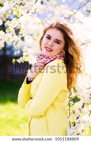A young woman dressed in a fashionable yellow coat in a flowering park. Stand sideways and smile.