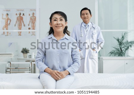 Pretty senior patient posing for photography with wide smile while sitting on bed of modern ward, highly professional doctor standing behind her