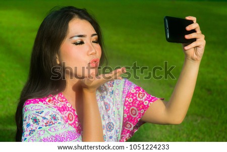 young happy and beautiful Asian Chinese woman taking selfie pic with mobile phone camera throwing a kiss posing outdoors on green grass park in internet social media addiction