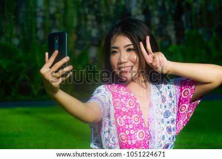 young happy and beautiful Asian Chinese woman taking selfie pic with mobile phone camera doing peace sign with hand fingers outdoors on green grass park in internet social media addiction