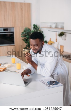 Smiling man doing online shopping and standing with credit card in kitchen