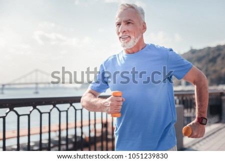 Sport with me. Confident senior unshaken man spending time on fresh air holding dumbbells and running. Royalty-Free Stock Photo #1051239830
