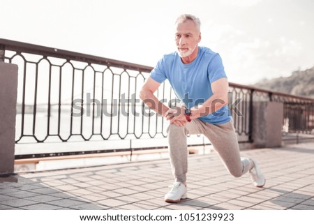 Sport forever. Senior confident unshaken making man exercises on the quay stretching legs and looking aside. Royalty-Free Stock Photo #1051239923