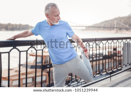 Warm up firstly. Concentrated unshaken strong man standing on the quay holding by the railings and stretching his leg. Royalty-Free Stock Photo #1051239944