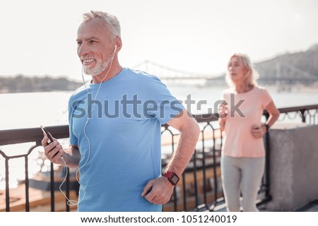 I like it. Satisfied unshaken senior man having morning training holding the smartphone and smiling. Royalty-Free Stock Photo #1051240019