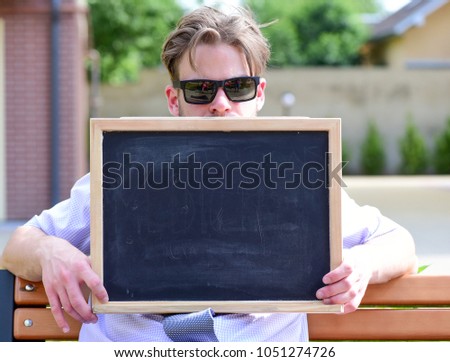 Salesman sits on bench with advertise for sale. Businessman with calm face with building on background. Business and real estate concept. Estate agent holds blackboard or chalkboard, copy space.