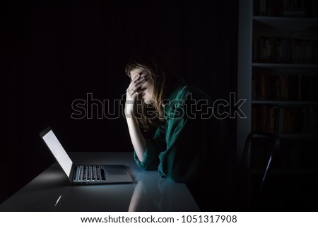 Tired and exhausted young woman hides eyes with hand at laptop pc late in the evening. Portrait of depressed female student or worker sitting in front of computer screen at night, concept of anxiety Royalty-Free Stock Photo #1051317908