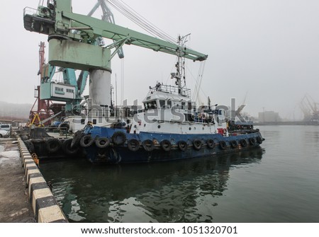 The tug is at the pier in the seaport. The background cranes shrouded in fog