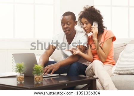 Young couple web surfing on laptop sitting on sofa at home, woman talking on phone, relaxing. Freelance, remote work and online shopping concept, copy space