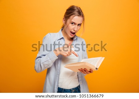 Image of displeased young lady student standing isolated over yellow background reading book. Looking camera.