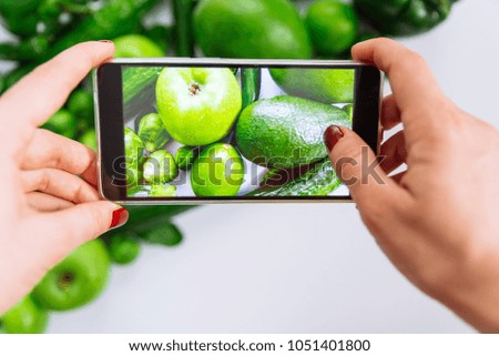 woman take picture of green vegetables and fruits on her phone. cucumber apple mango lime on white background
