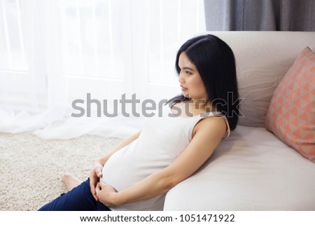 Beautiful young Asian pregnant woman sits by the window and touches her tummy. Pregnancy. Health. Motherhood. A cozy portrait of a pregnant woman.